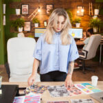 Young woman reviewing print-on-demand designs at desk with laptop and printed samples.