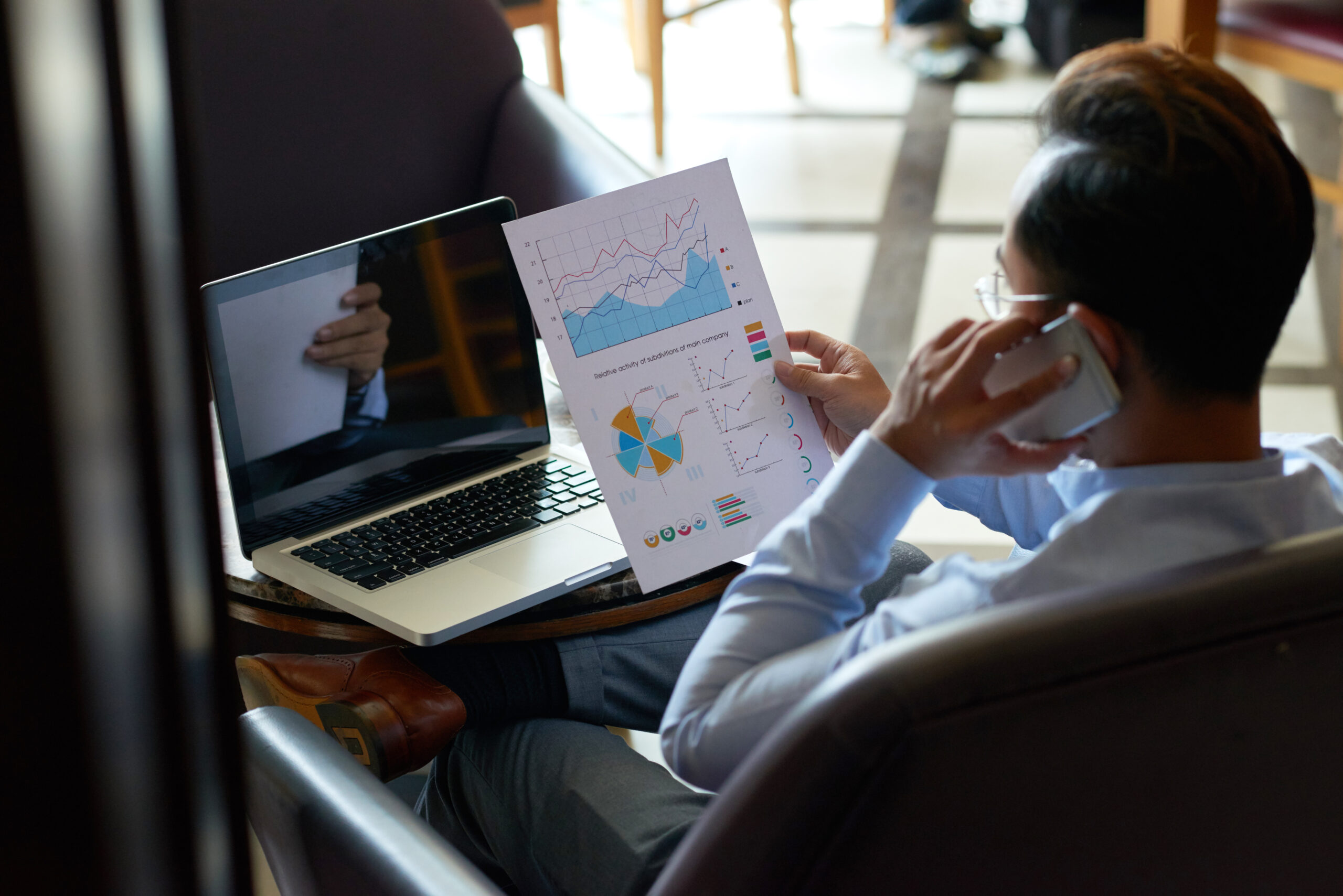 Man multitasking by reviewing financial documents and having a telephone chat while conducting market research for his online business.